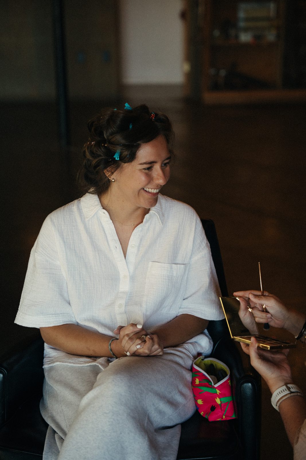 Bride in a white linen set with hair curlers laughing while someone holds a makeup palette near her