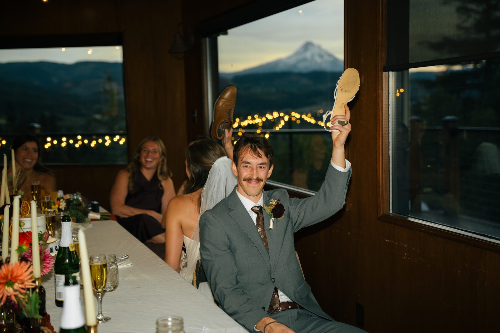 Groom raising a shoe at the head table during the shoe game, Mount Hood glowing in the window behind him