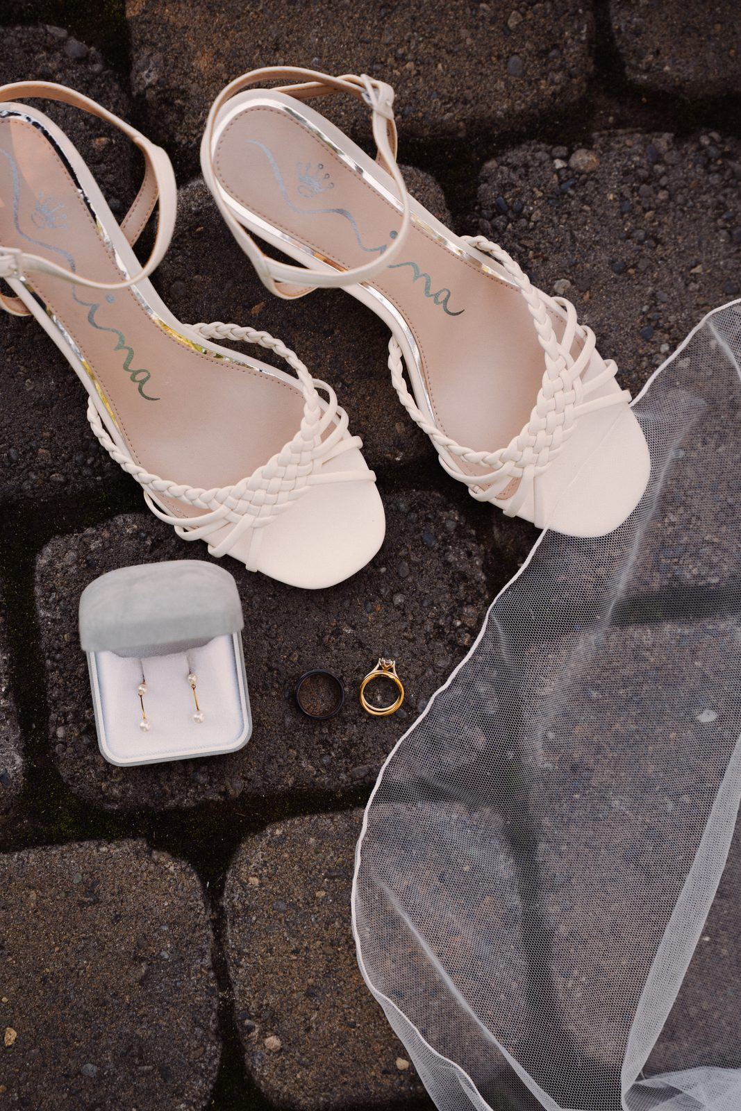Braided white wedding sandals, pearl earrings in a box, two wedding rings and a veil laid out on stone pavers