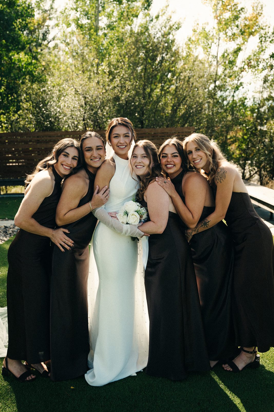 Becca in the middle of her five bridesmaids in brown satin dresses, all hugging in the sun