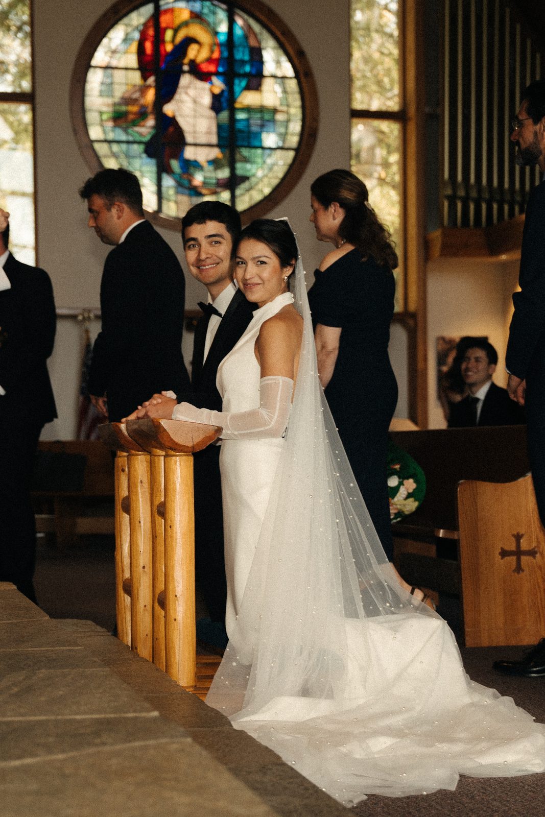 Skyler and Becca smiling back at the camera while kneeling at the altar rail, with a stained glass window behind them