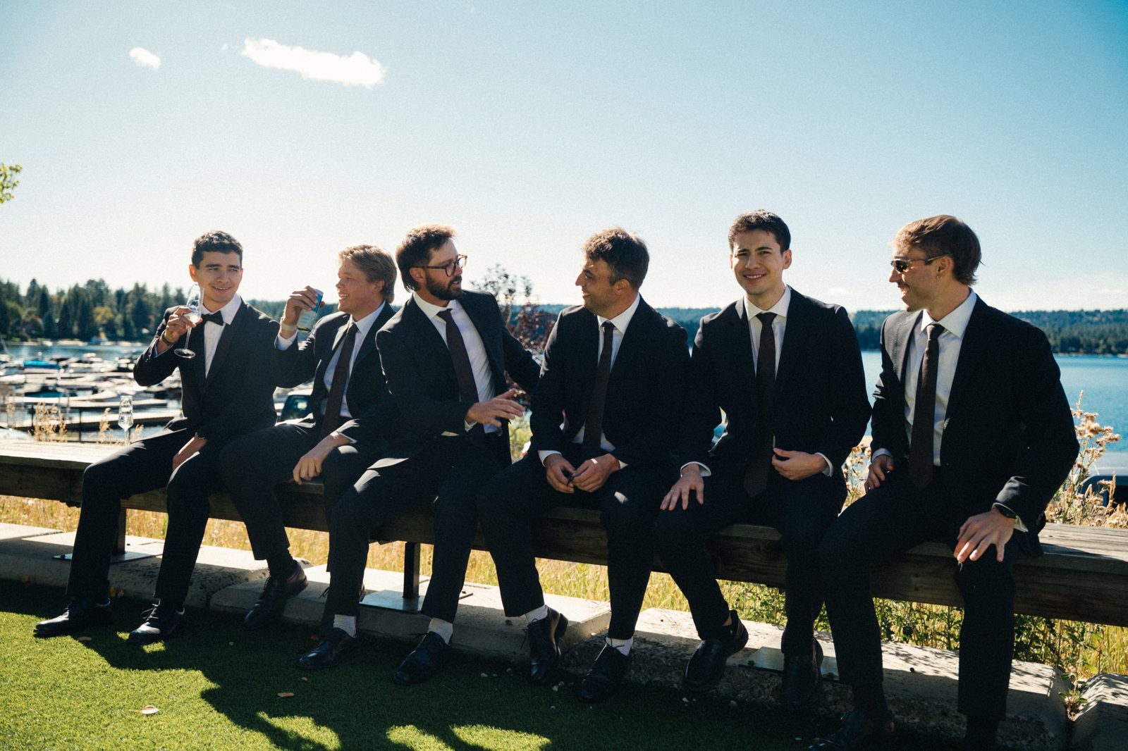Skyler and his groomsmen in black suits laughing and joking on a wooden bench beside the lake