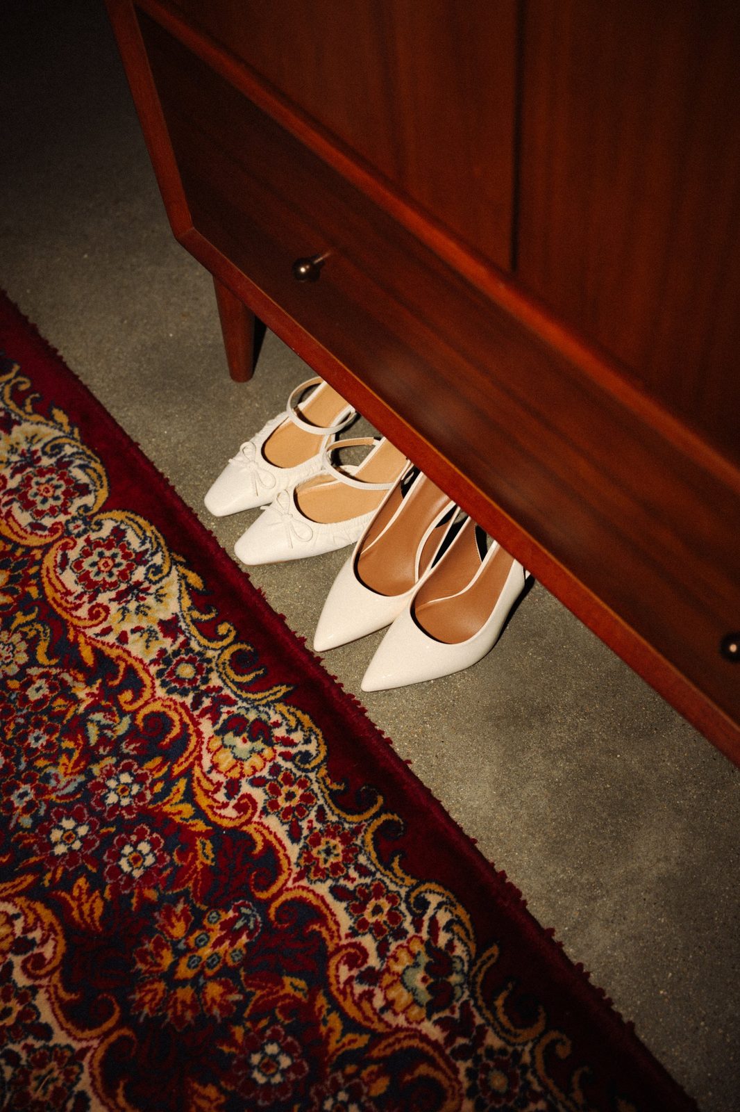Becca’s white wedding shoes tucked under a wooden dresser next to a red patterned rug