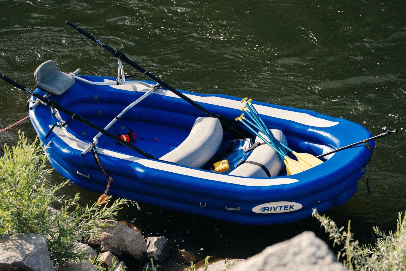 A blue RIVTEK raft with oars, seats, paddles and a dry box tied up against a rocky river bank, seen from above