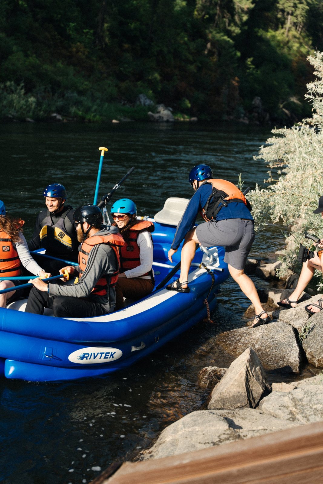 A guide stepping onto a blue RIVTEK raft from the rocks while guests in helmets and life jackets wait, as a videographer films from the shore