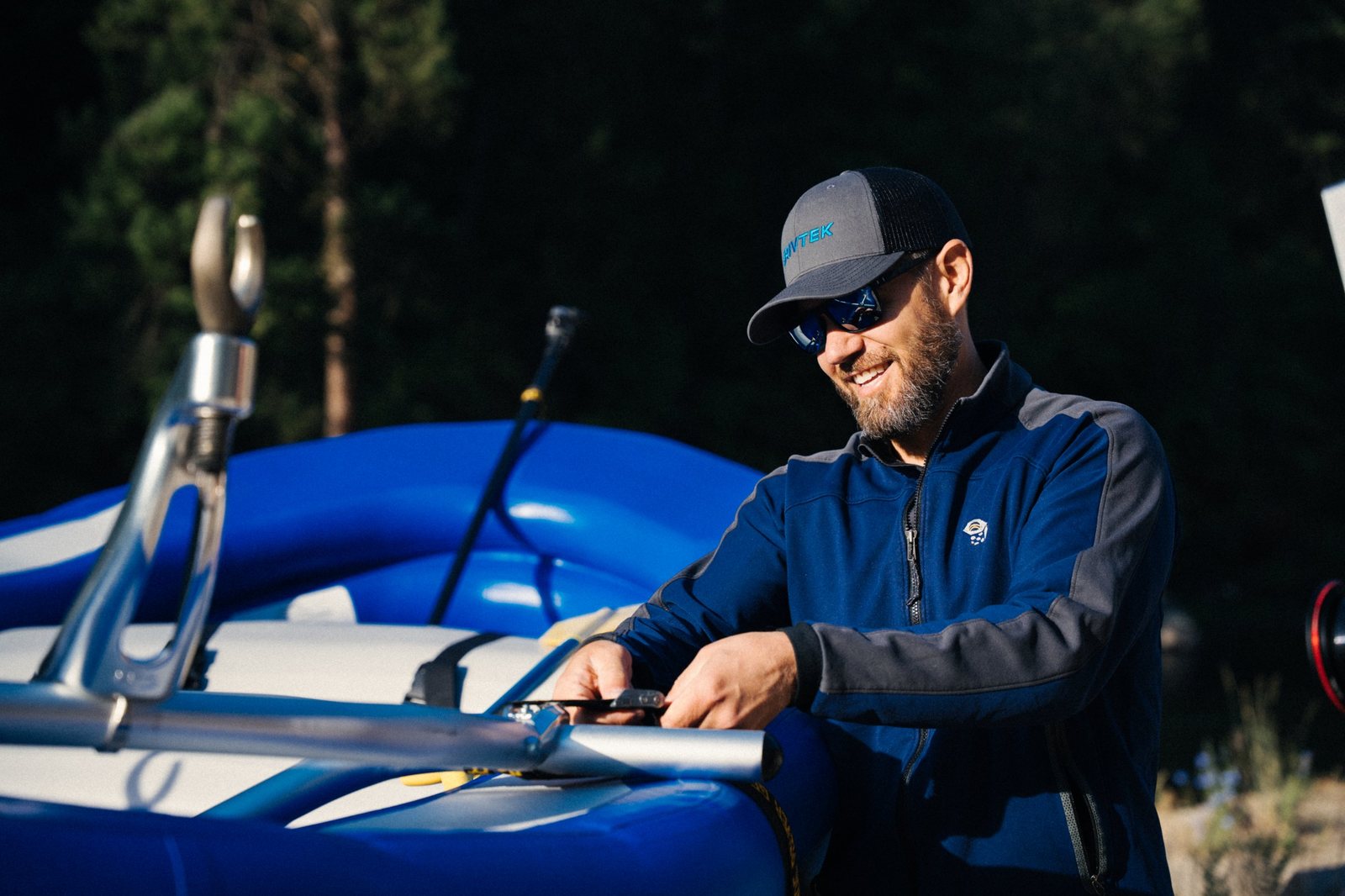 A smiling bearded man in sunglasses and a RIVTEK cap cinching a strap on a blue raft with a chrome oar mount in the foreground