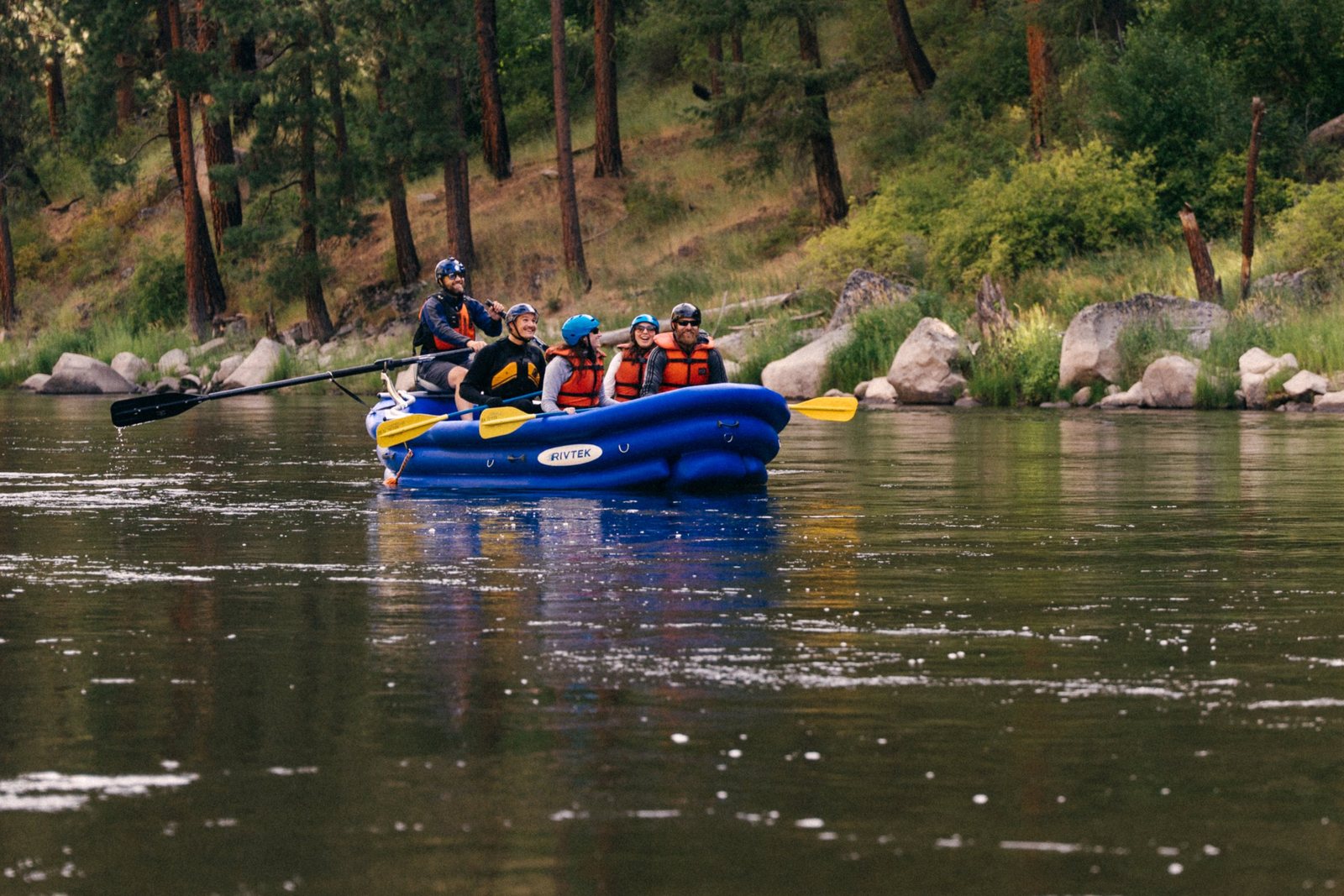 A blue RIVTEK raft with five smiling people in helmets and life jackets floating on a calm river below a pine-covered hillside