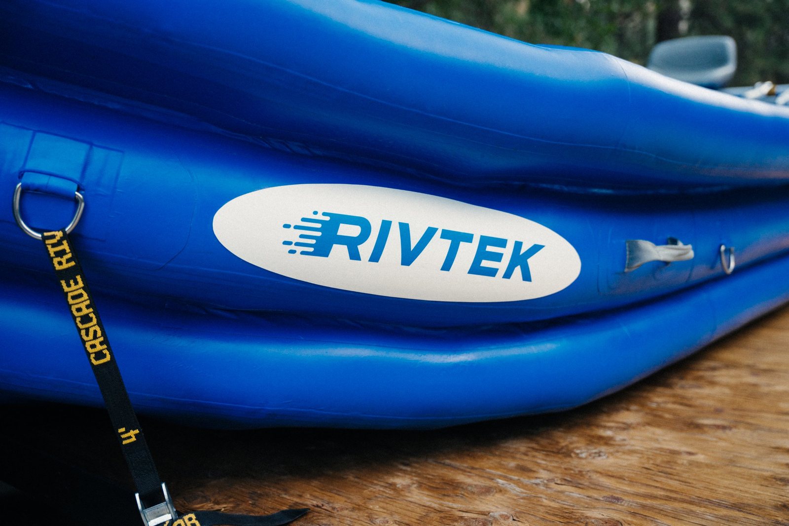 Close-up of the RIVTEK logo on the side of a blue inflatable raft, with a D-ring and a black Cascade River strap in the foreground