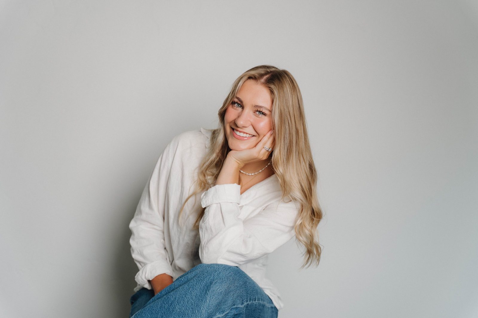 A smiling woman with long blonde hair resting her chin on her hand in a white linen shirt against a light gray wall