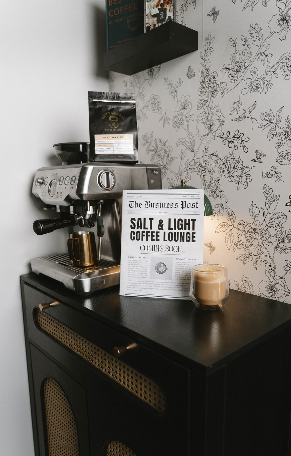 A coffee corner with an espresso machine, a Kettlebell coffee bag, a Salt & Light Coming Soon newspaper and a latte on a black cane cabinet