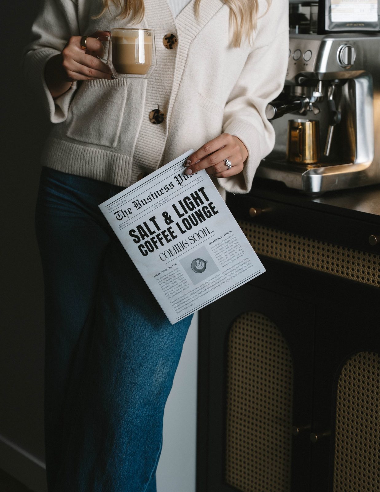 A Salt & Light Coffee Lounge Coming Soon newspaper on a dark counter beside a green mushroom lamp and an espresso machine, with floral wallpaper behind