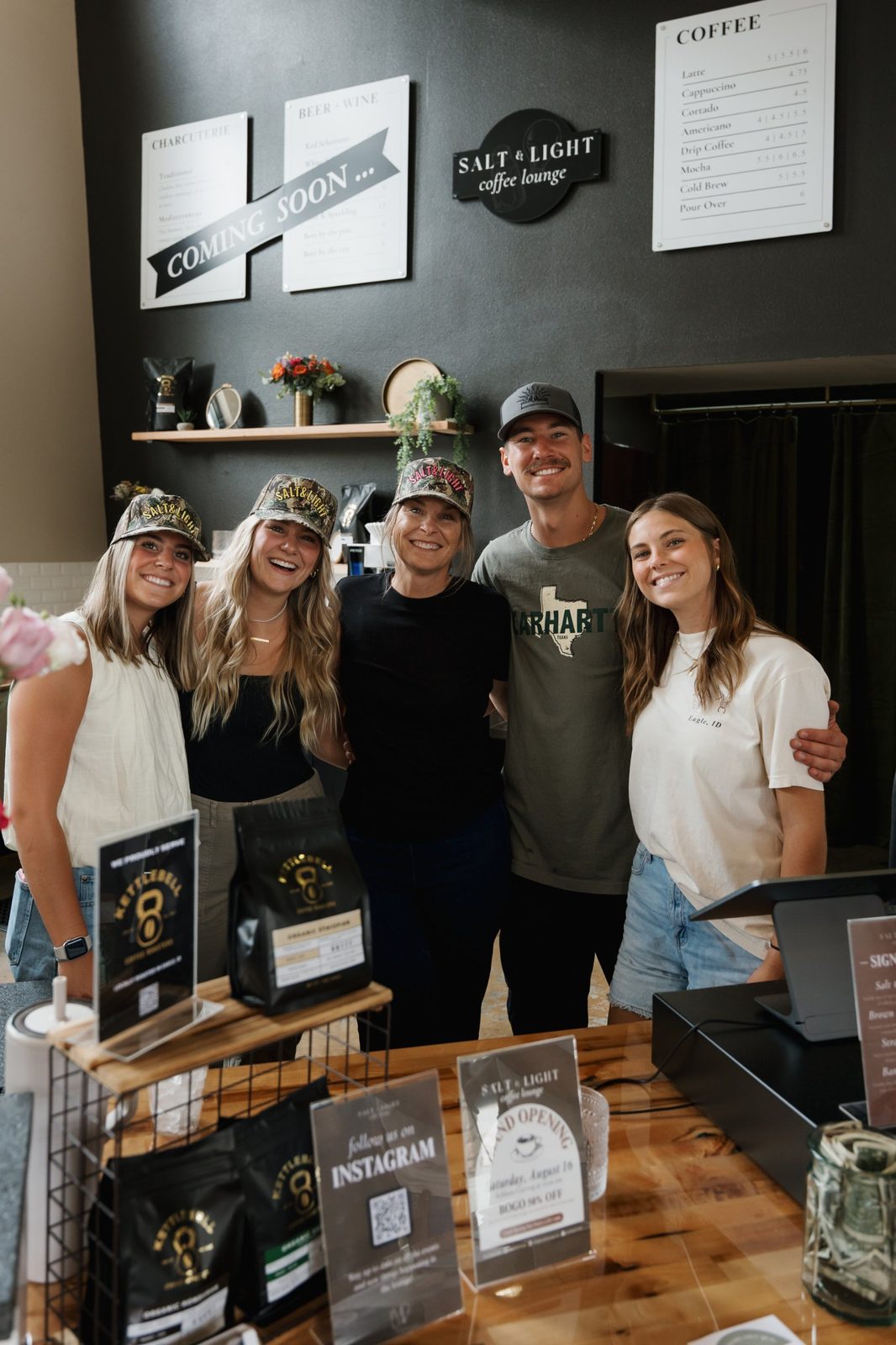 Five smiling Salt & Light team members, most in camo caps, behind the coffee counter on grand opening day with menu boards and a Coming Soon banner behind them