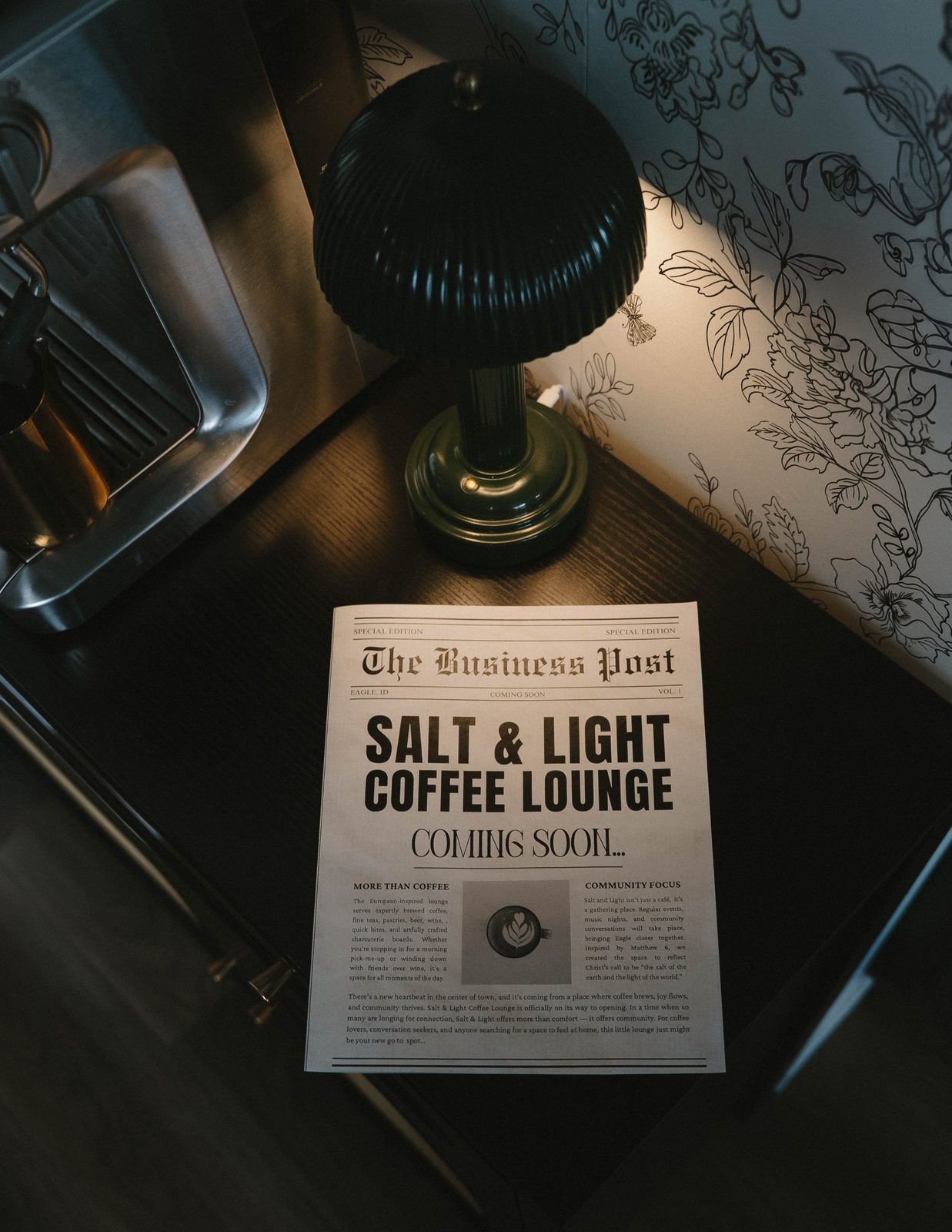 Close-up of a woman in a cream cardigan holding a latte in one hand and a Salt & Light Coming Soon newspaper in the other, leaning on a cane cabinet