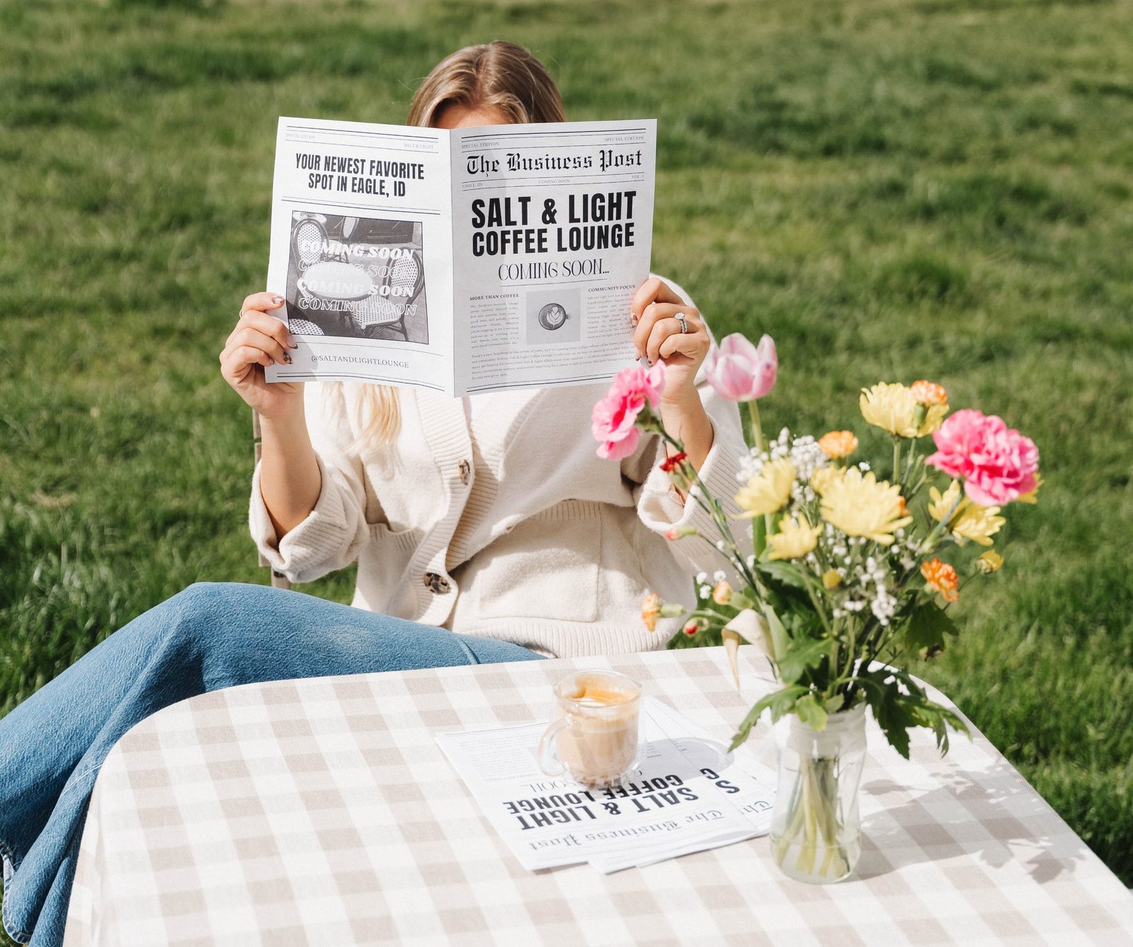 A woman hiding behind an open Salt & Light Coming Soon newspaper at a gingham-covered table with a latte and a bouquet of spring flowers on the grass