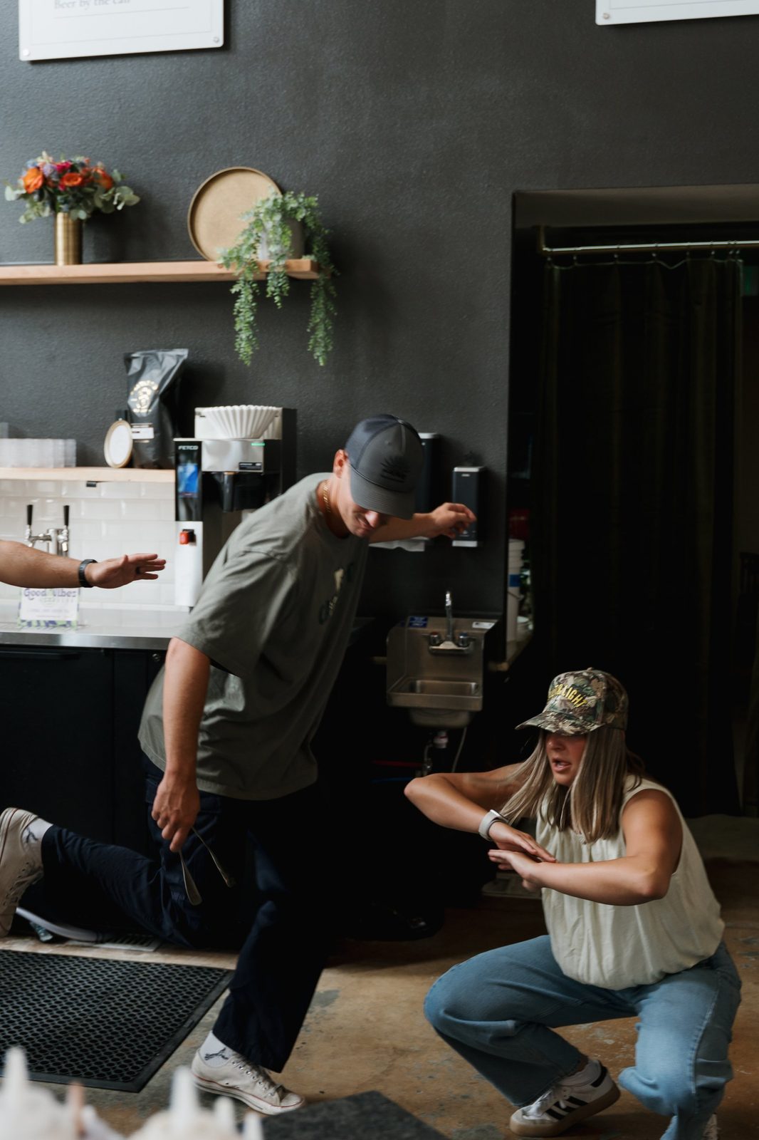 A man in a gray cap and a woman in a camo Salt & Light cap striking playful poses behind the coffee bar