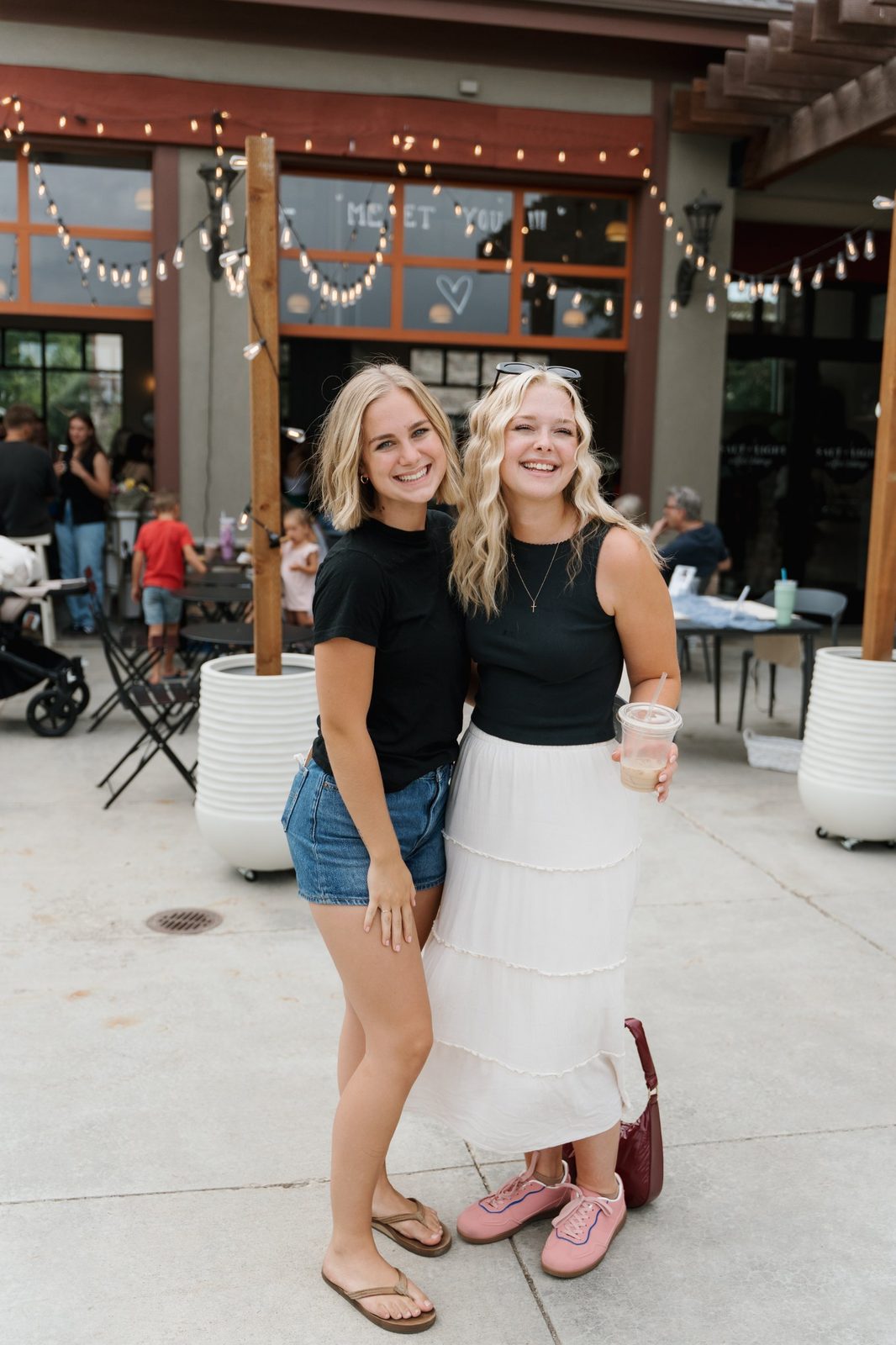 Two smiling friends posing on the patio outside Salt & Light under string lights, one holding an iced coffee