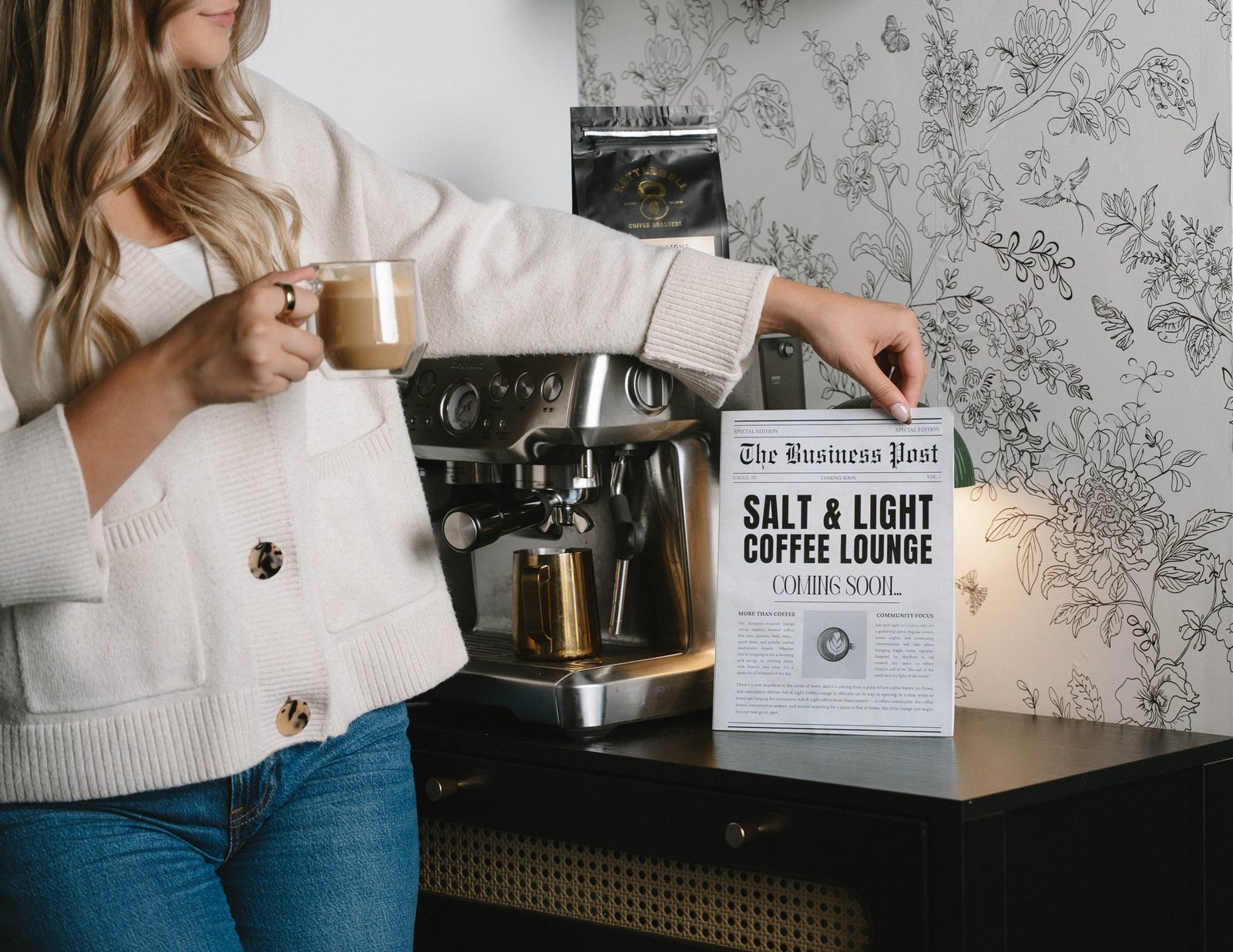 A woman in a cream cardigan holding a latte and resting her other hand on a Salt & Light Coffee Lounge Coming Soon newspaper beside an espresso machine and floral wallpaper