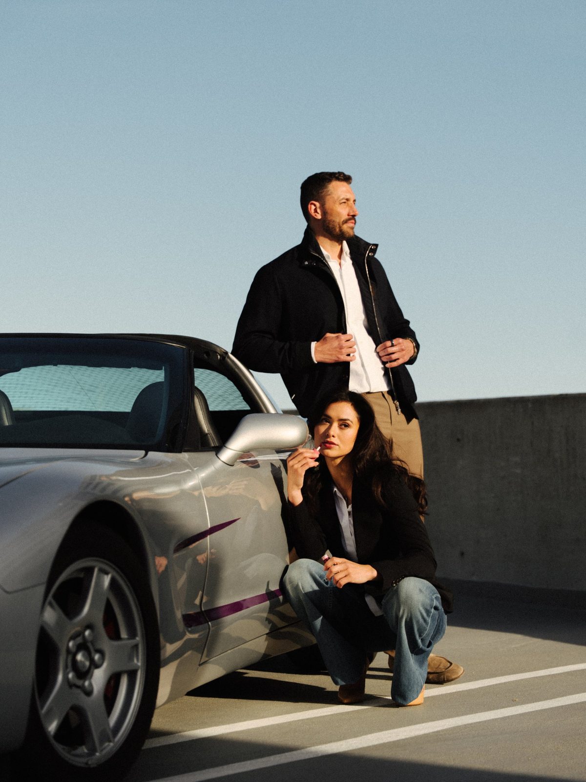 Man in a navy jacket standing beside a silver convertible while a woman crouches next to it applying lip gloss, rooftop brand shoot in Boise, Idaho