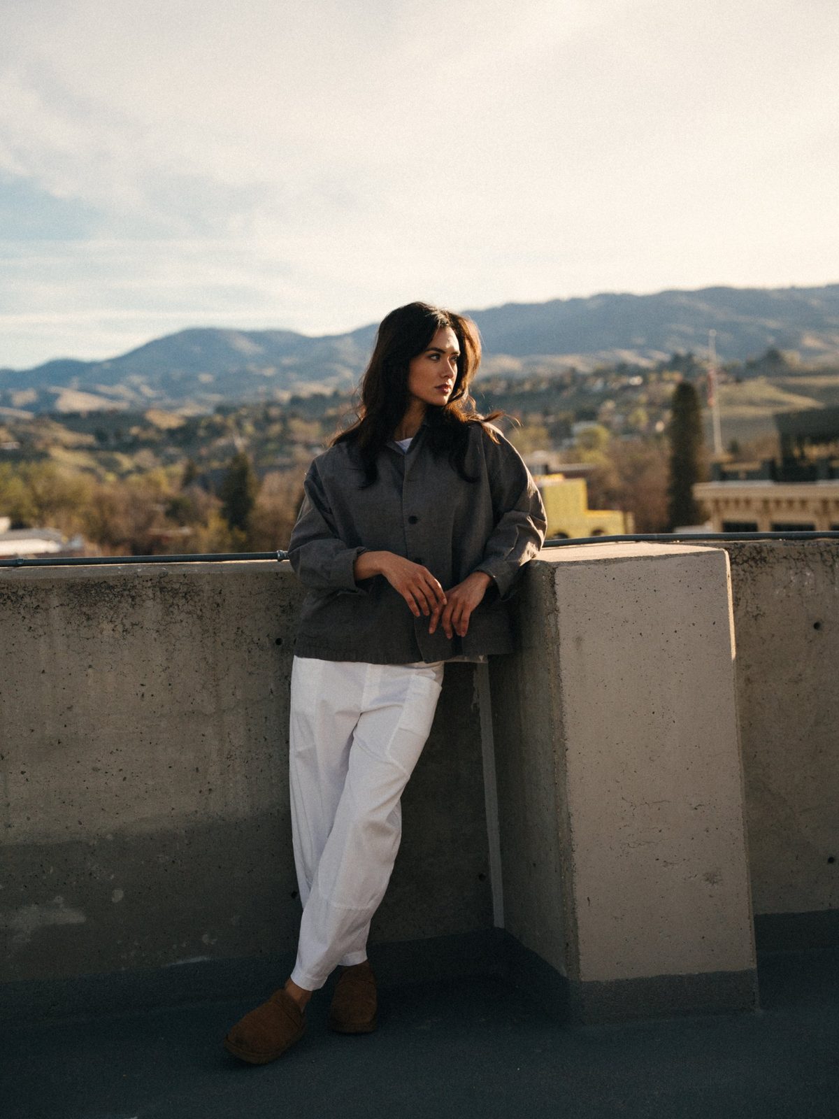 Woman in a gray linen jacket and white trousers leaning on a rooftop wall with the Boise foothills behind her