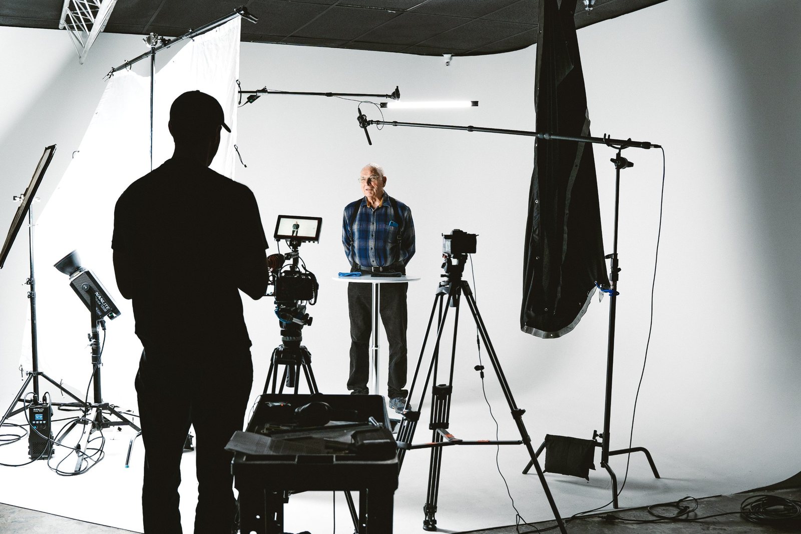 Behind the scenes on a white studio set: a camera operator in silhouette frames an older man standing at a small table, with lights, boom mics and tripods around them