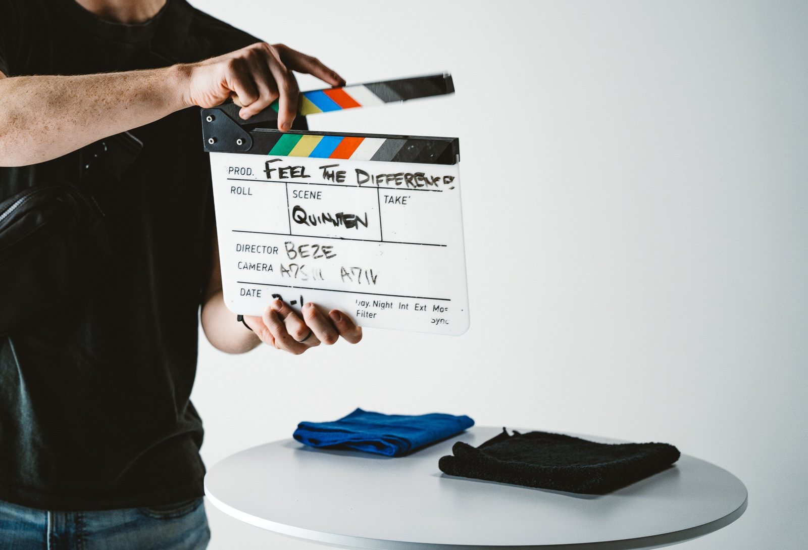 A hand holding a clapperboard marked Feel the Difference in front of a white round table with a folded blue towel and a folded black towel