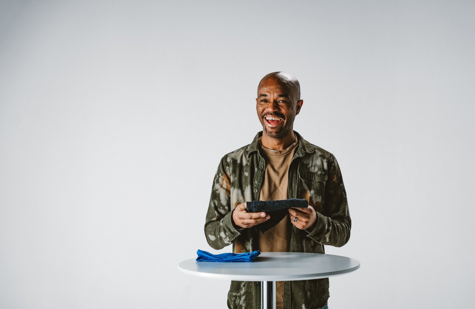 A smiling man in an olive green jacket holding a folded black towel at a white standing table with a blue towel, against a light gray studio wall