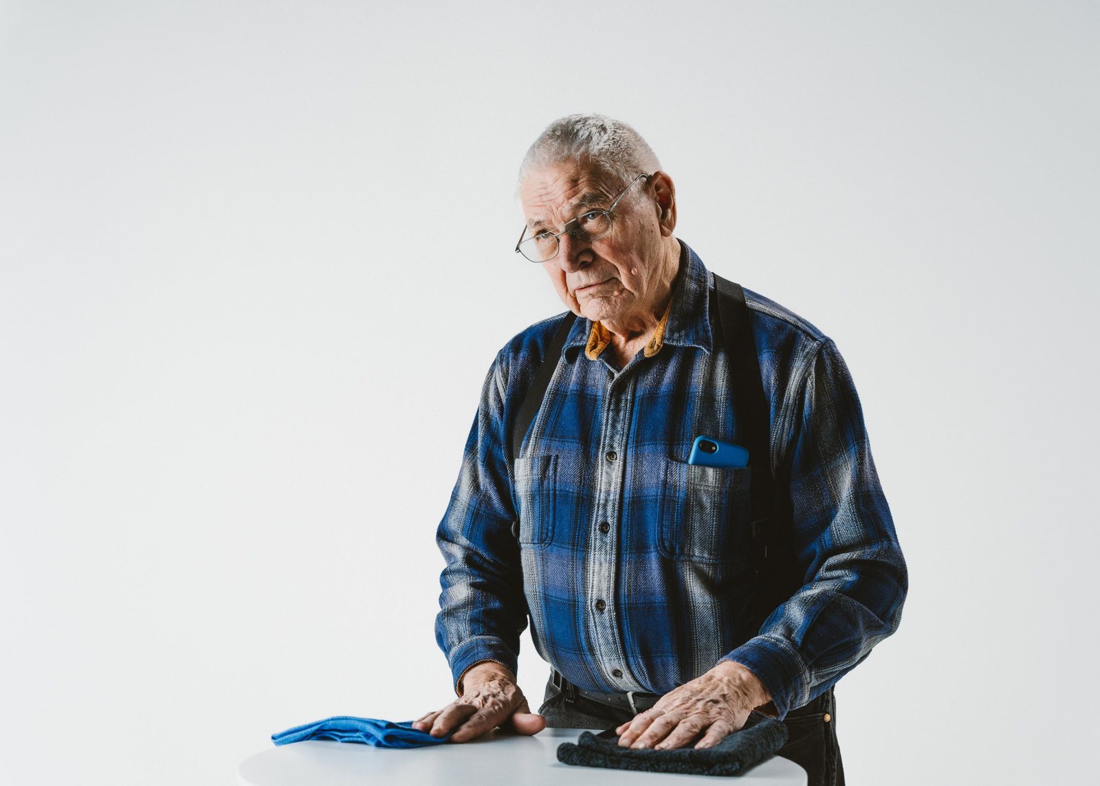 An older man with gray hair and glasses in a blue plaid flannel shirt and suspenders resting his hands on a white table with two folded towels, against a clean white studio backdrop