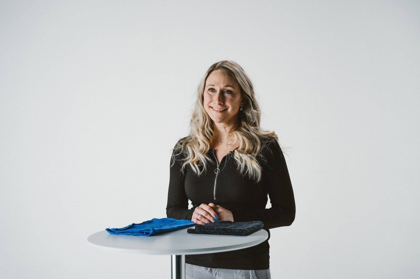 A woman with long blonde hair in a black zip-up top smiling at a white standing table with a blue towel and a black towel, against a white studio backdrop